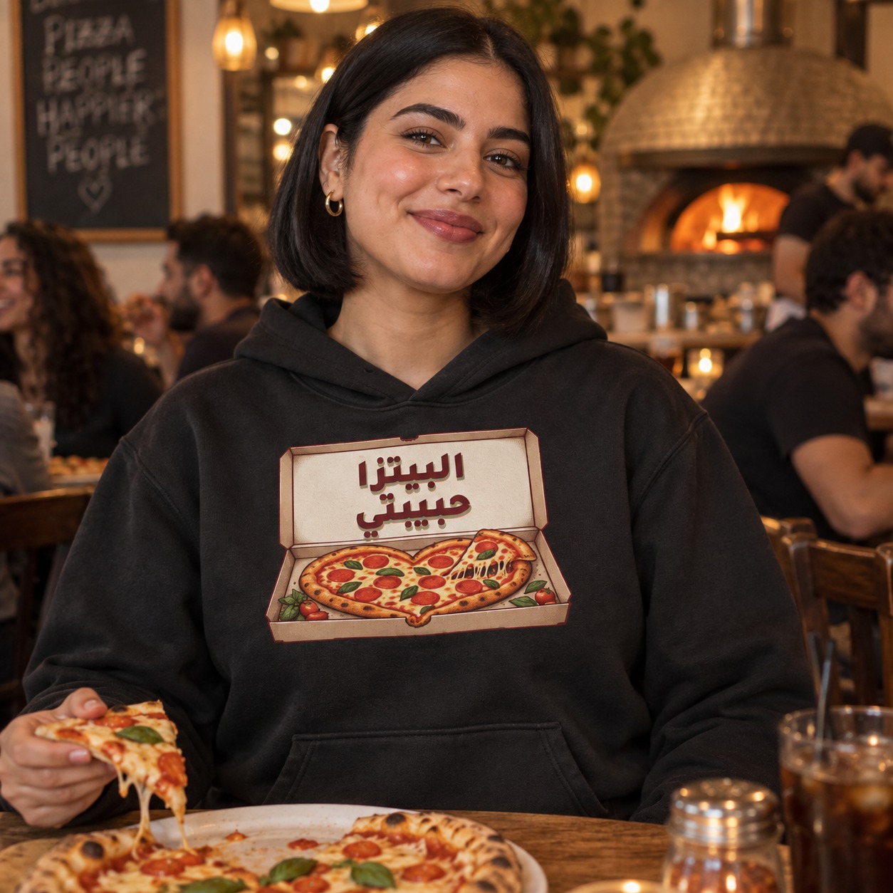هودي أسود بيتزا ذهبية على شكل قلب وقطعة مرفوعة بخيوط جبنة داخل علبة مفتوحة تحمل البيتزا حبيبتي بخط خمري وظل أخضر هادئ